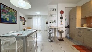 Una cocina moderna con armarios blancos, un comedor con mesa de vidrio y una sala de estar con sofá y un cuadro.