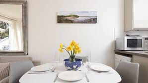 Un coin repas avec une table dressée pour deux personnes, un vase bleu contenant des fleurs jaunes et un tableau accroché au mur.