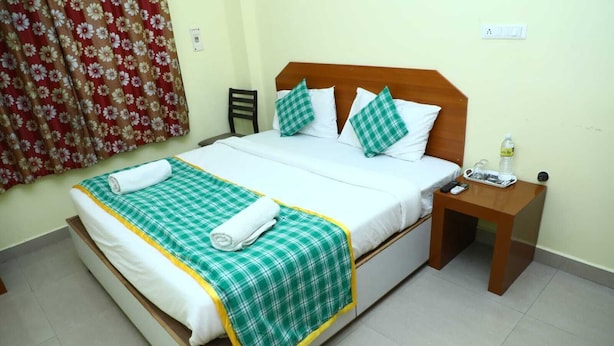 A neatly made bed with a green and white checkered bedspread, two pillows, and a rolled white towel. A small wooden nightstand with a bottle and remote control is beside the bed. - Shiva Nest (Tiruchirappalli)