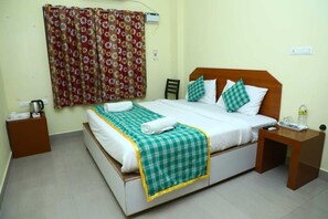 A neatly arranged hotel room with a bed, a bedside table, a nightstand, a window with curtains, and a wall socket. - Shiva Nest (Tiruchirappalli)