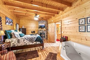 A log cabin bedroom with a bed, a fireplace, a stone wall, and a bathroom with a bathtub.