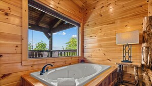 A wooden cabin interior with a large bathtub, wooden walls, a window with a view, and a small table with a lamp. - Long range sunset views from Hot Tub, free attractions, Resort pool (Sevierville)