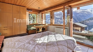 A bedroom with a large bed, wooden walls, a window with a mountain view, and a small table with chairs.