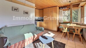 A cozy living room with a sofa, a small table, and a dining area with chairs. There is a kitchen area with cabinets and a window providing natural light.