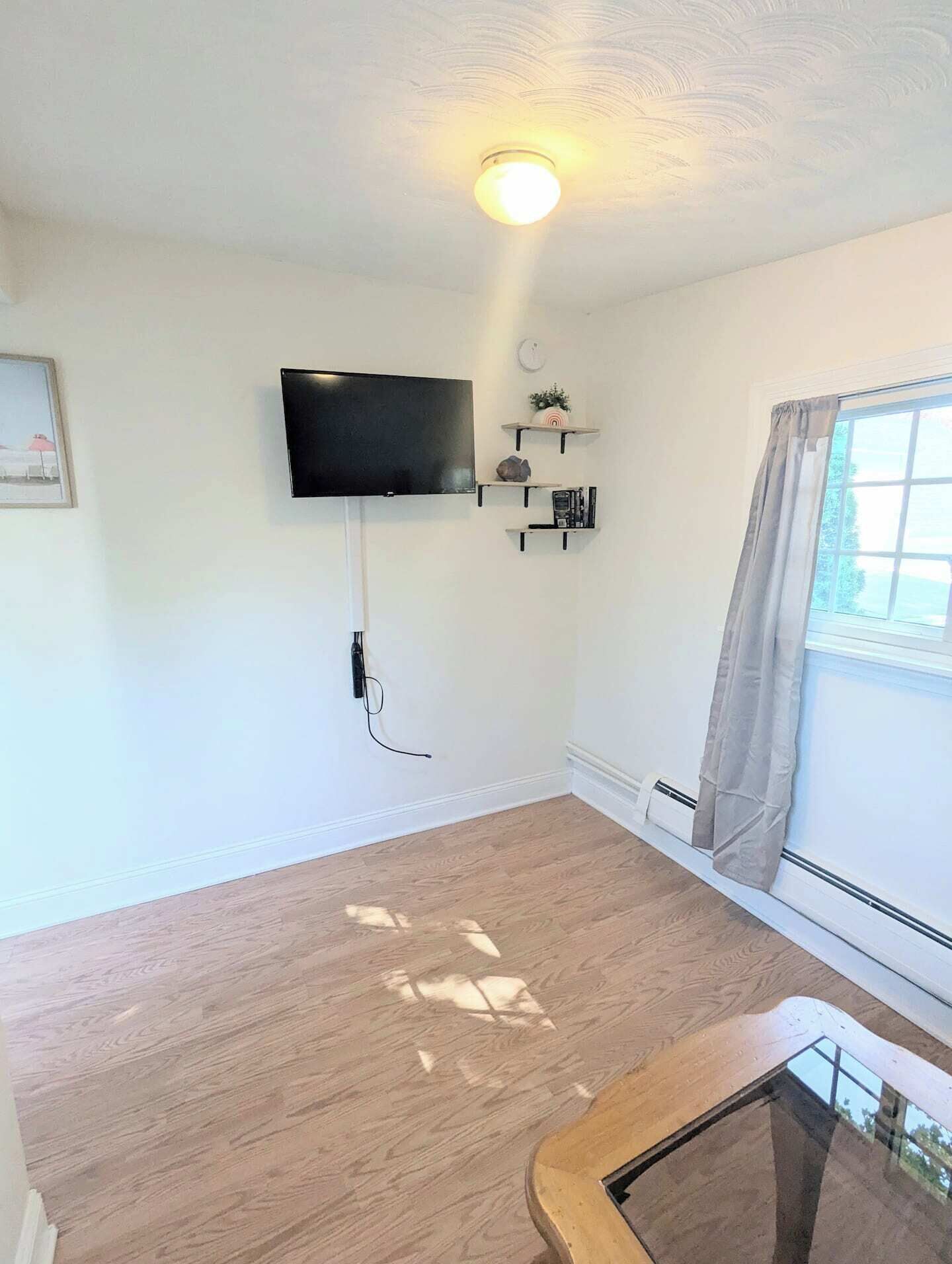 Een kamer met een wandgemonteerde flatscreen televisie, een klein plankje met decoratieve objecten, een raam met gordijnen en een houten vloer.