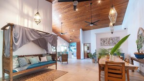 A spacious living area with a wooden ceiling, a wooden sofa with a canopy, a dining table with chairs, and a kitchen in the background.