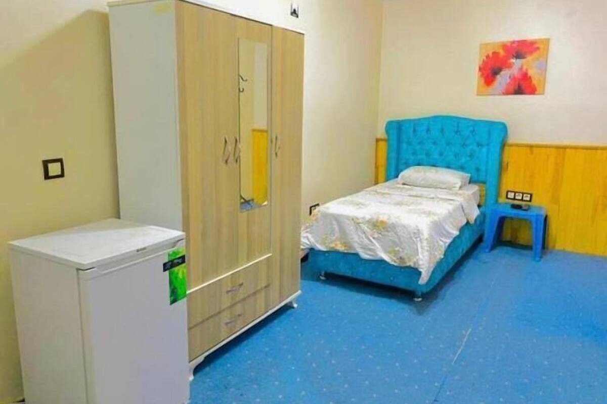A bedroom with a bed, a wardrobe, a small table, and a wall painting.