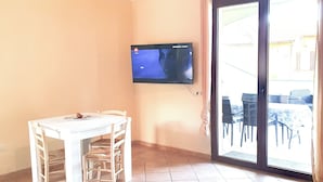 A room with a dining table and chairs, a flat-screen TV mounted on the wall, and a sliding glass door leading to an outdoor area. - Bilocale Confort Viviana-LaCiaccia Style (La Ciaccia)