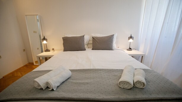 Een netjes opgemaakt bed met twee kussens, een grijze deken en twee opgerolde handdoeken. Er staan twee nachtkastjes met lampen bij.
