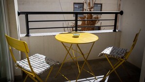 Een balkon met een gele tafel en twee gele stoelen, een kleine schaal op de tafel en een uitzicht op gebouwen achterin.