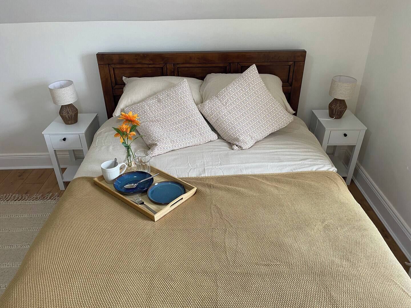 Un lit bien fait, avec une tête de lit en bois, deux tables de chevet avec des lampes, et un plateau contenant des assiettes et un vase de fleurs posé sur le lit.