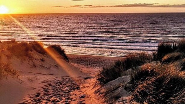 A beach scene with the sun setting over the ocean, sandy dunes, and a clear sky.