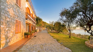 A stone pathway leading to a building with a balcony overlooking a grassy area and a body of water. - Il Giardino dei Filosofi (Ascea)