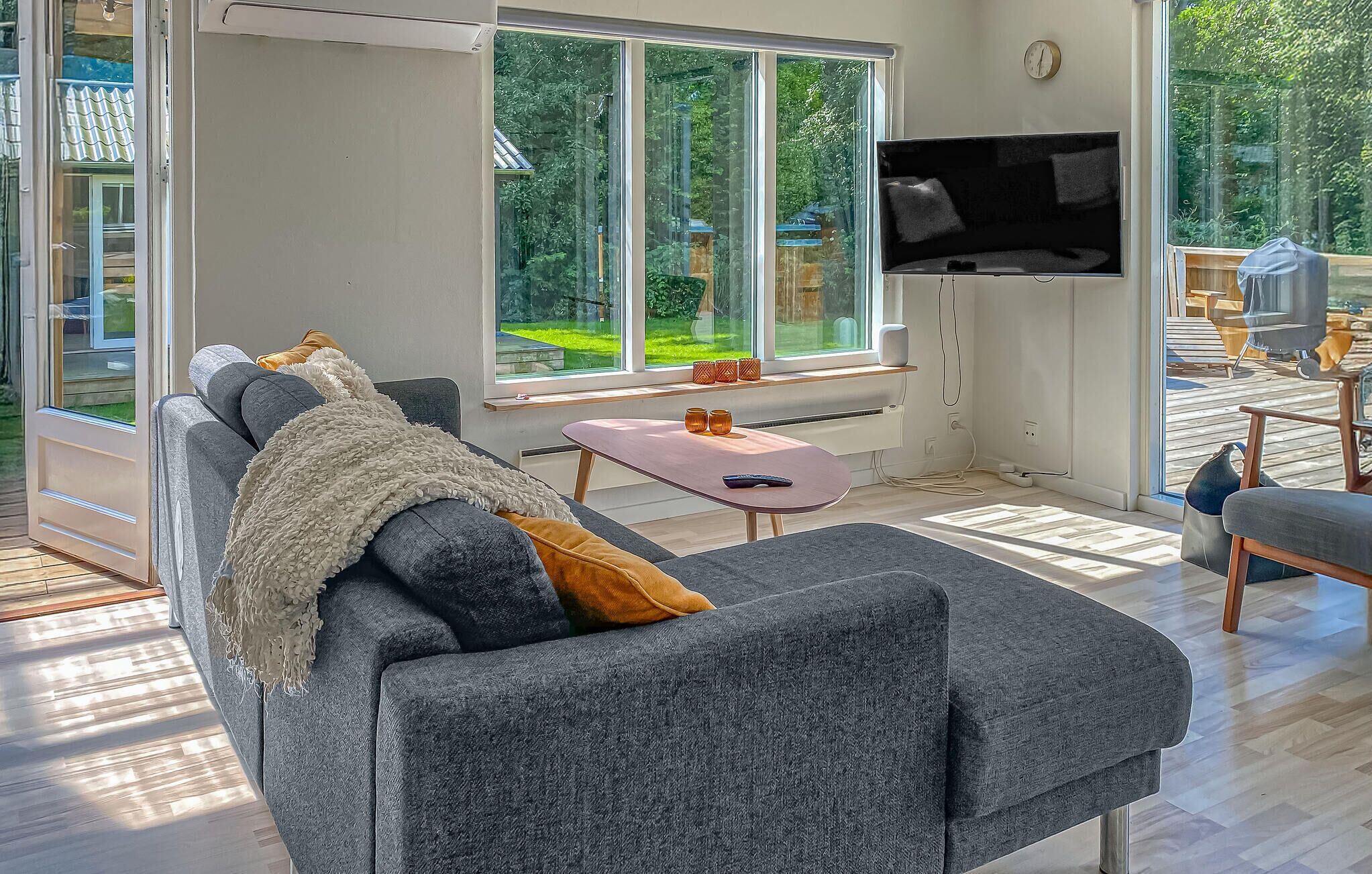 A modern living room with a grey sofa, a flat-screen TV, and a large window overlooking a garden.