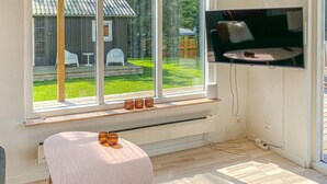 A modern living room with a large window, a flat-screen TV mounted on the wall, a round table with a remote control, and a small shelf with decorative items. - Gorgeous home in Odder with WiFi (Odder)