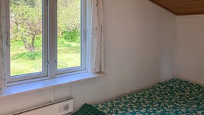 Et soveværelse med et vindue, en radiator, en seng med blomstret dyne og udsigt til grøn vegetation udenfor.