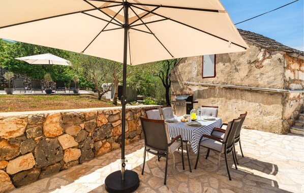 Un espace extérieur aménagé pour les repas, avec des murs en pierre, une table dressée pour deux personnes et un barbecue à l’arrière-plan.