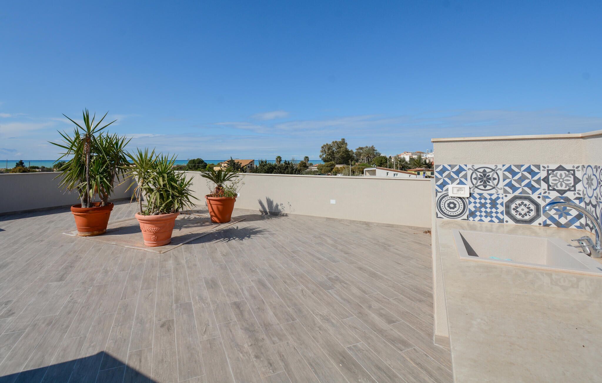 A rooftop terrace with three potted plants, a tiled wall with decorative patterns, and a view of the sea and buildings in the distance.