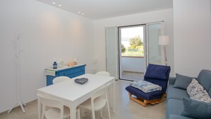 Ein modernes Wohnzimmer mit einem weißen Tisch, einem blauen Sofa und einem blauen Sessel. Eine Glastür führt zu einem Balkon mit Blick auf Grünflächen.