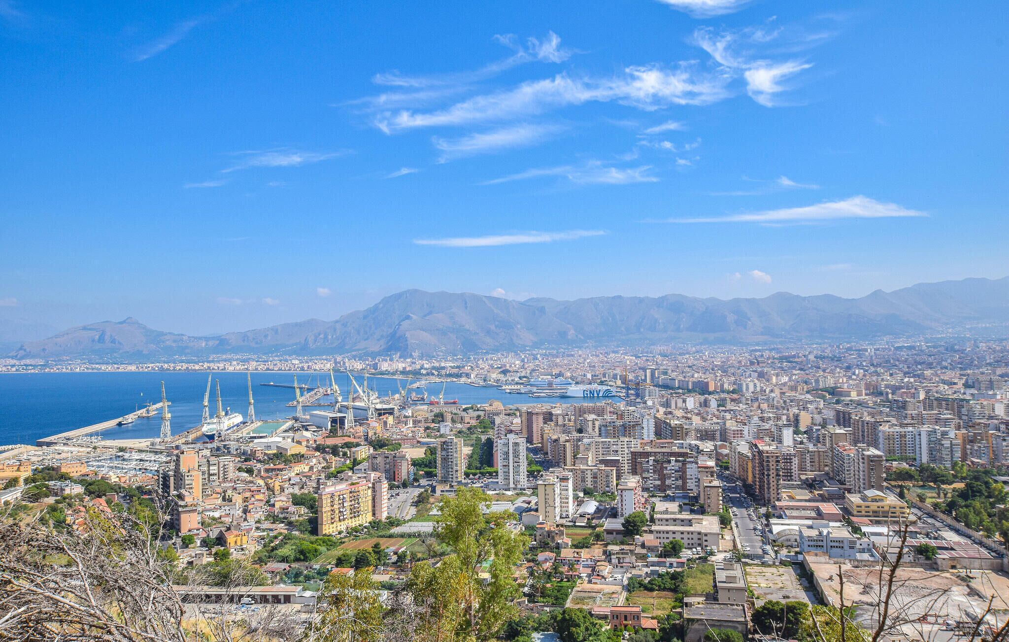 A coastal cityscape with a marina, high-rise buildings, and mountains in the background.
