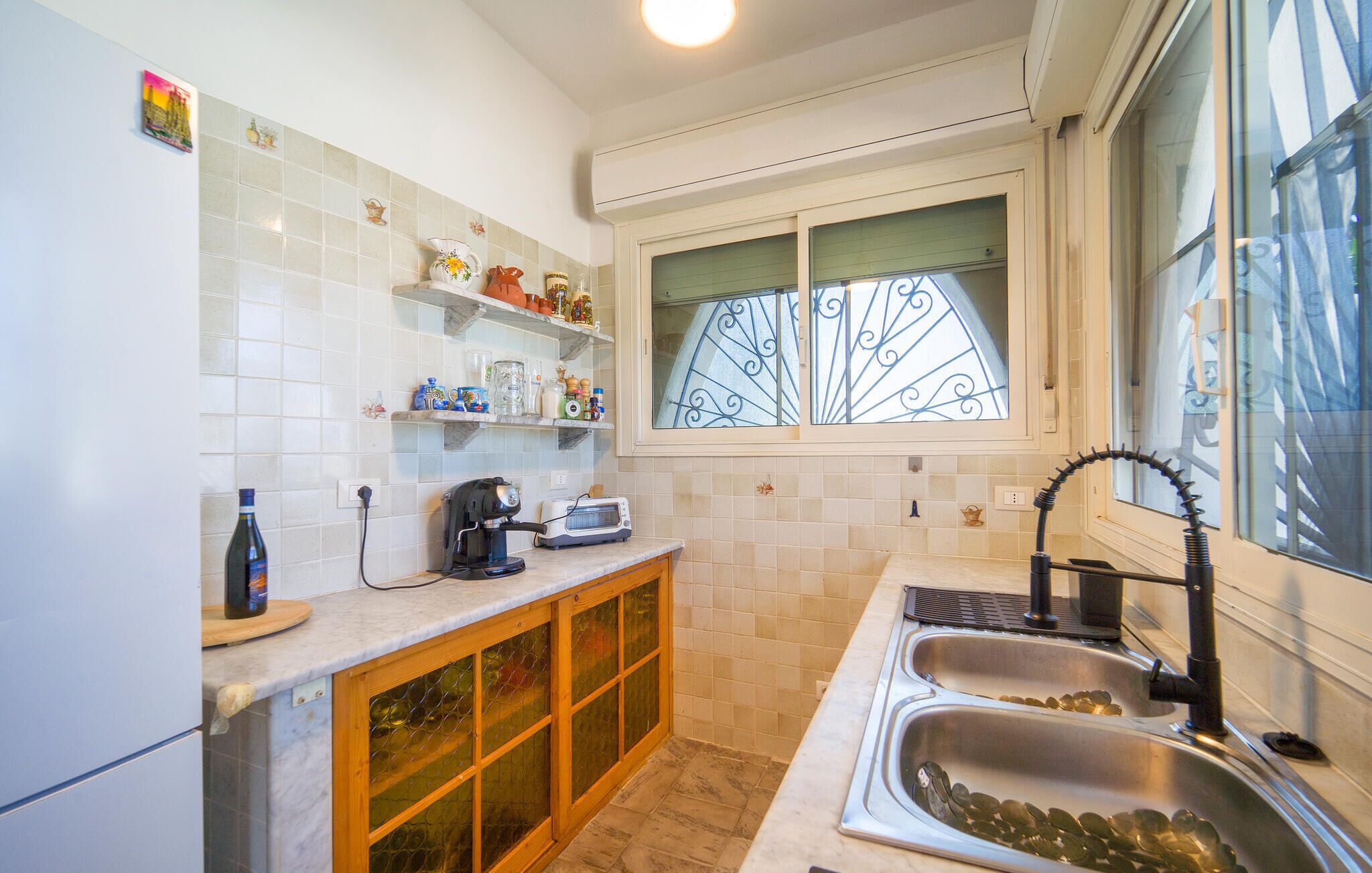 A compact kitchen with white tiles, a window with decorative iron bars, a built-in oven, and a sink with a faucet.