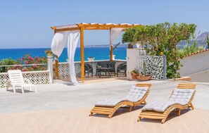 Uma área de estar ao ar livre ensolarada com espreguiçadeiras e um pérgola, com vista para um mar azul e límpido.