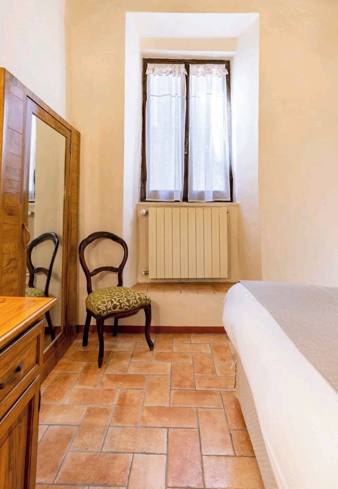 Une petite chambre avec une commode en bois, une chaise avec un coussin à motifs, un lit, une fenêtre avec des rideaux légers et un radiateur.