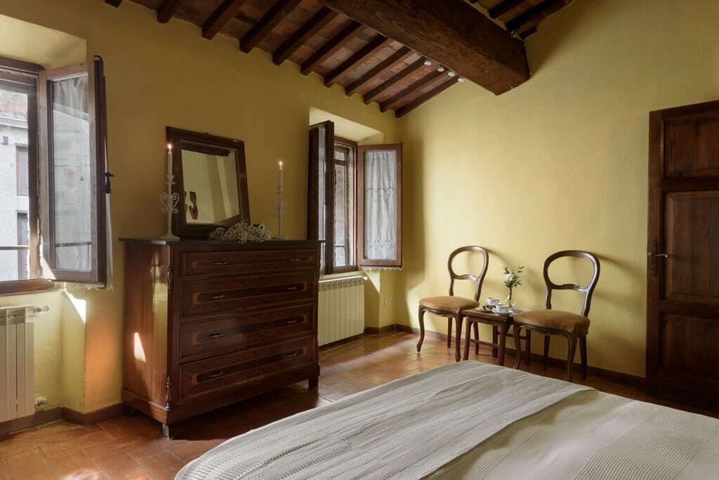 Un dormitorio con cómoda de madera, cama con cubrecama blanco, dos sillas, una mesa con un florero y una ventana con cortinas.