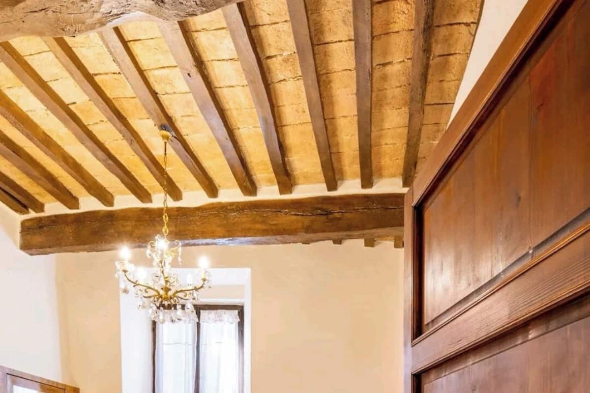 Un dormitori amb sostre de fusta, llit amb roba de llit blanca, un penjallum, una finestra amb cortines i un radiador.