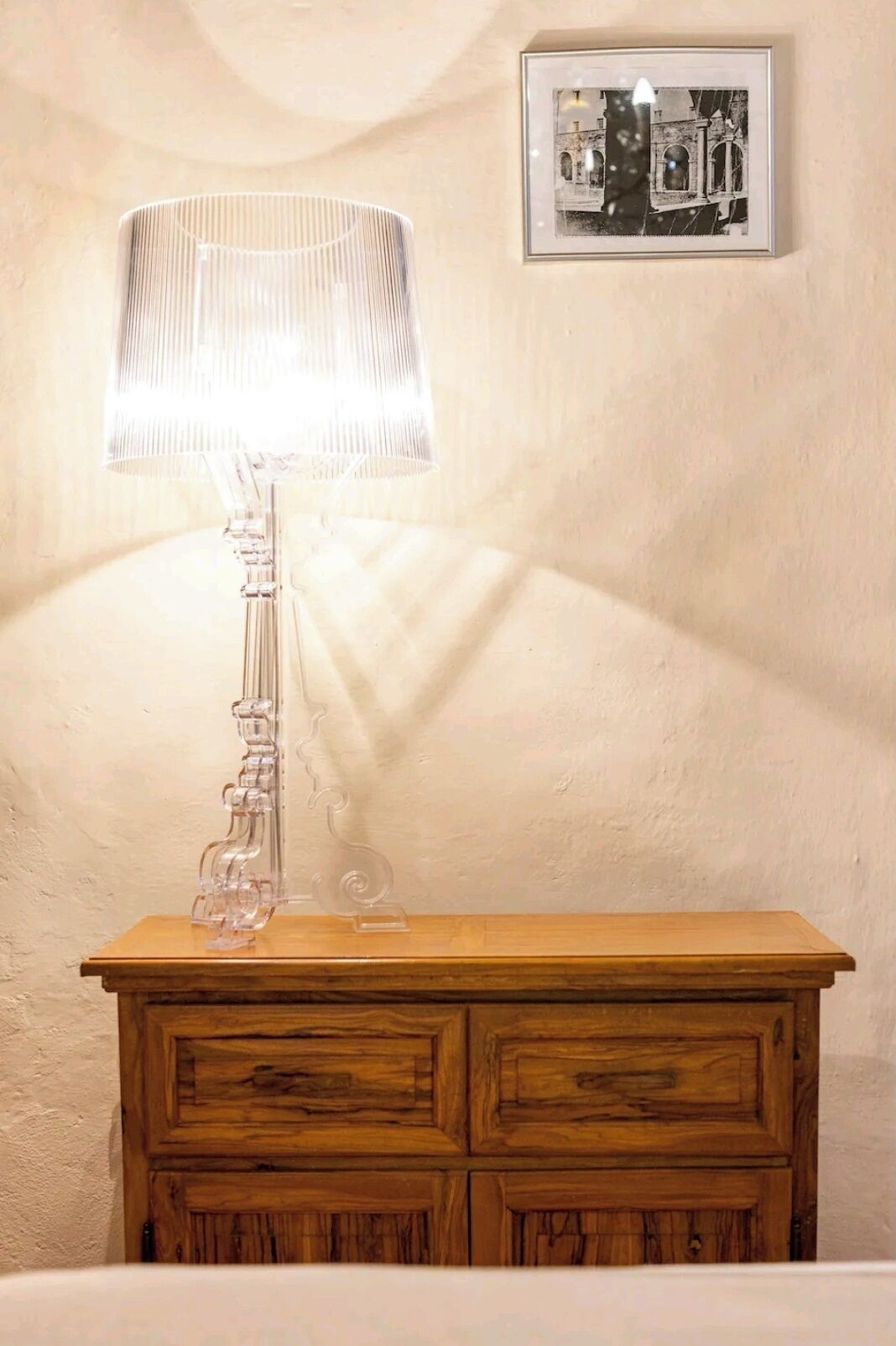 Une table de chevet en bois, sur laquelle se trouve une lampe à abat-jour transparent et, au mur, une photographie en noir et blanc encadrée.