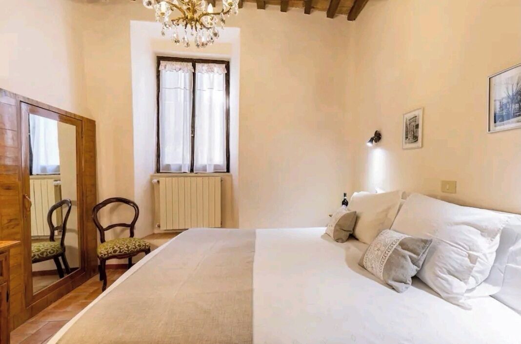 A bedroom with a large bed, a wooden wardrobe, a chair, a window with curtains, and a chandelier.