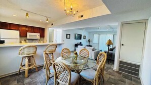 A modern kitchen with a dining area, featuring wicker chairs, a glass table, and a white refrigerator. - Bay Tree cozy 2BR condo on the ground floor (Little River)