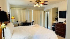 Habitación de hotel con una cama grande, ventilador de techo, mesa de comedor con sillas y televisor montado en la pared.
