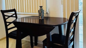 Un comedor con una mesa redonda y cuatro sillas, un florero sobre la mesa, y persianas verticales en las ventanas.