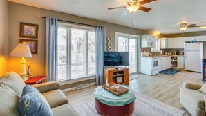 A living room with a sofa, a coffee table, and a television. There is a kitchen area with cabinets and a refrigerator.