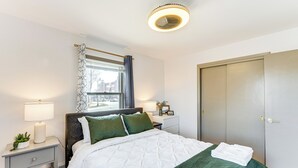 A neatly made bed with a white and green quilt, a bedside lamp, a small plant, and a window with a curtain.