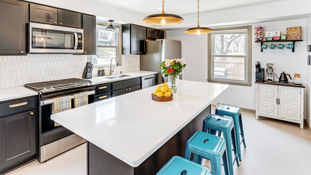 A modern kitchen with dark cabinets, a white island, blue bar stools, and a window with a view.