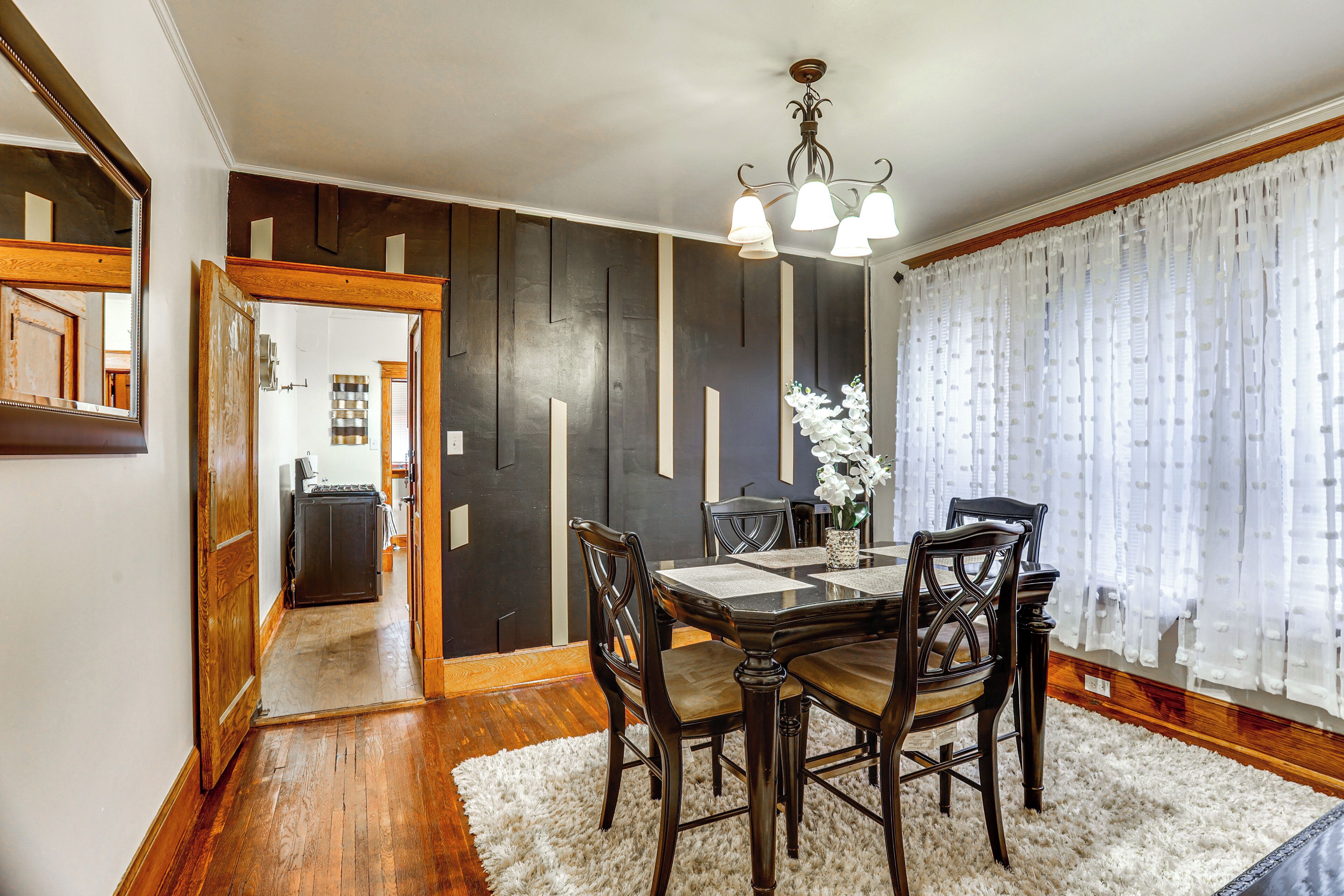 A dining area with a wooden table, chairs, and a chandelier. There is a white rug on the floor and a window with sheer curtains.