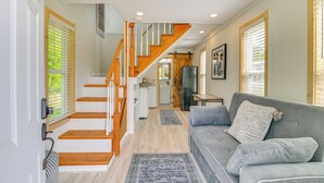 A spacious living area with a grey sofa, wooden staircase, and a kitchen in the background.