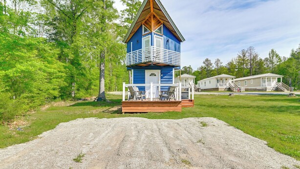A blue cabin with a wooden deck and a porch, surrounded by trees and a gravel path.