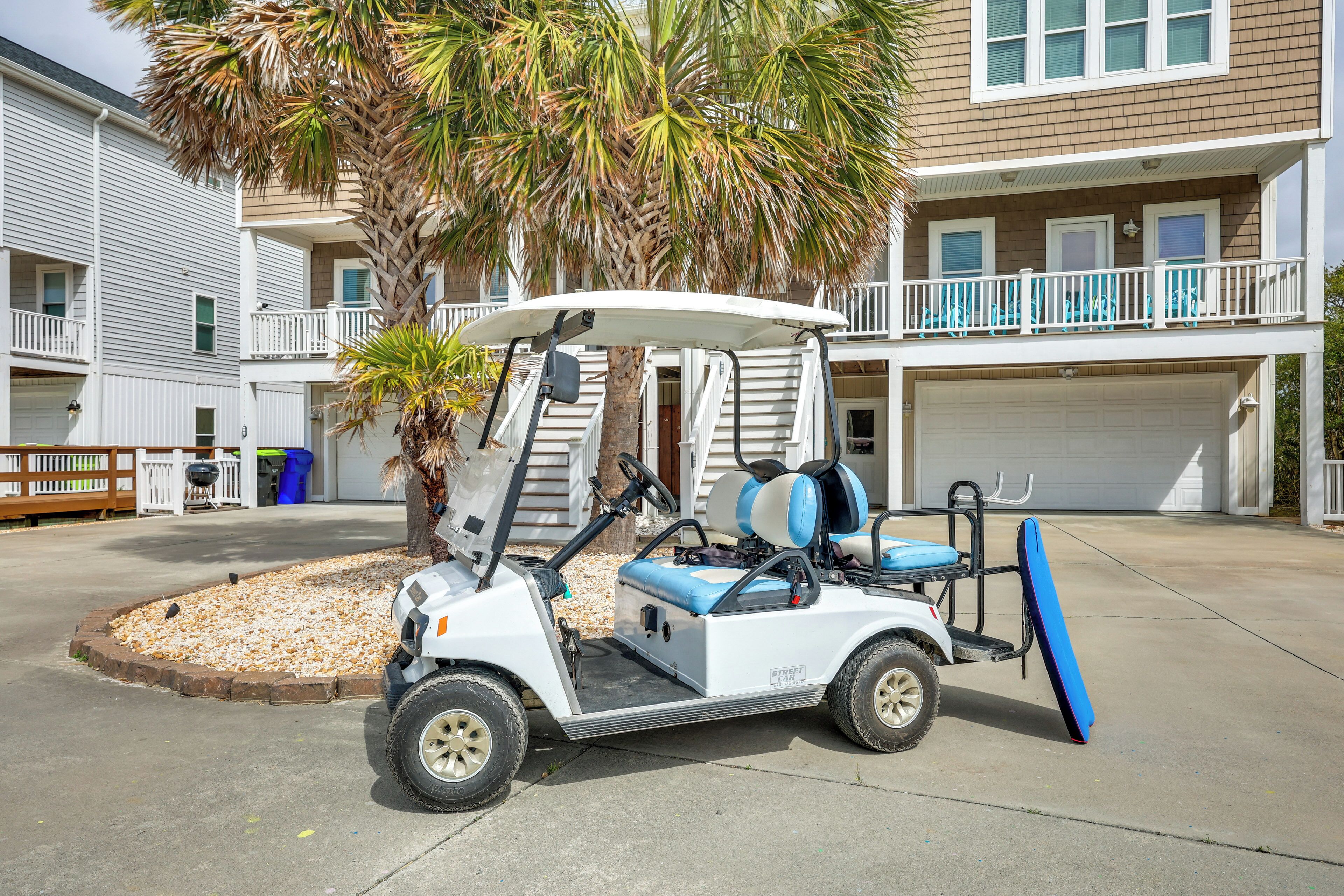A golf cart with a blue seat and a surfboard attached, parked in front of a building with a white exterior and balconies.
