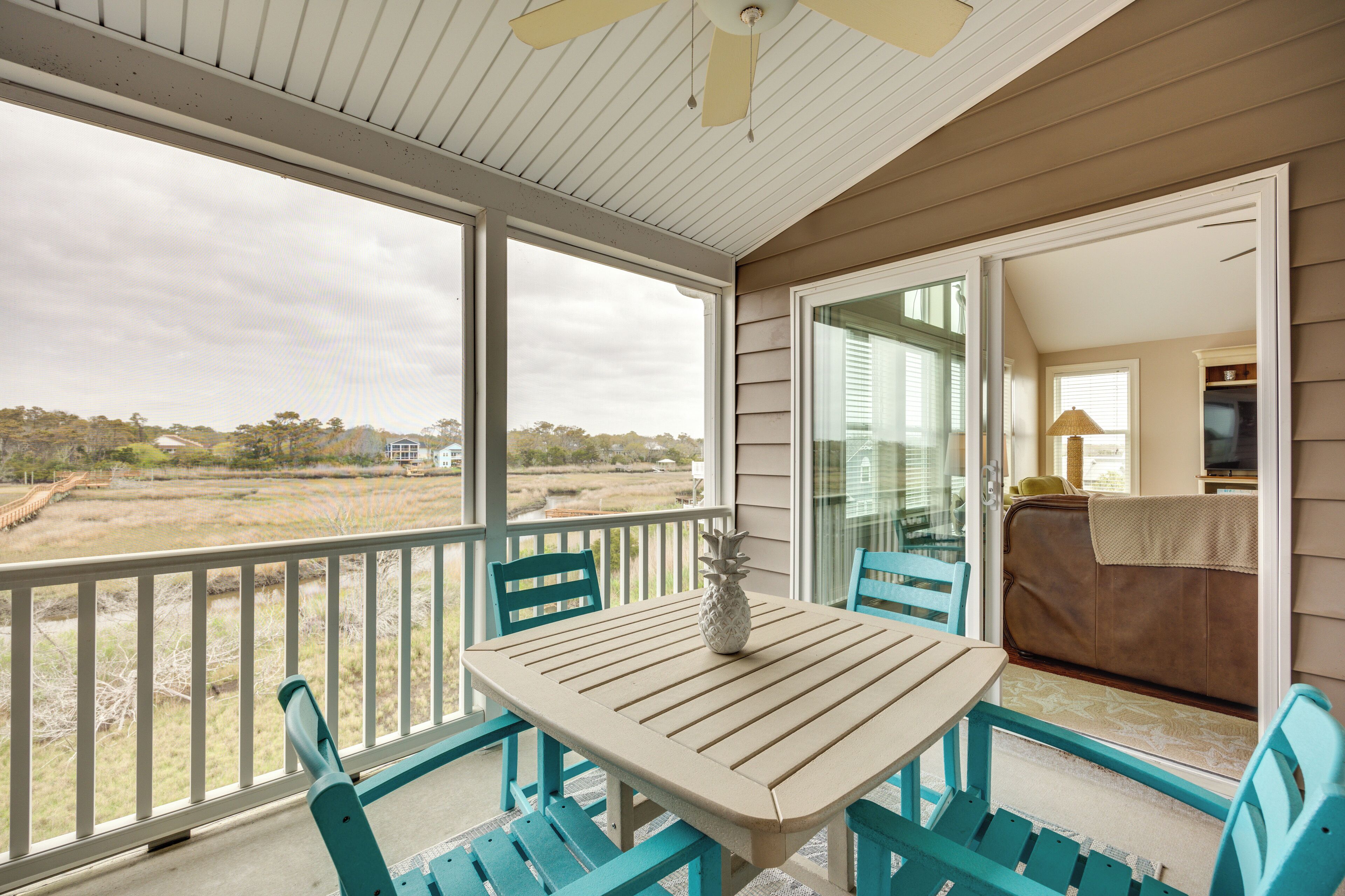 A covered balcony with a table and chairs, overlooking a beach and a distant house.