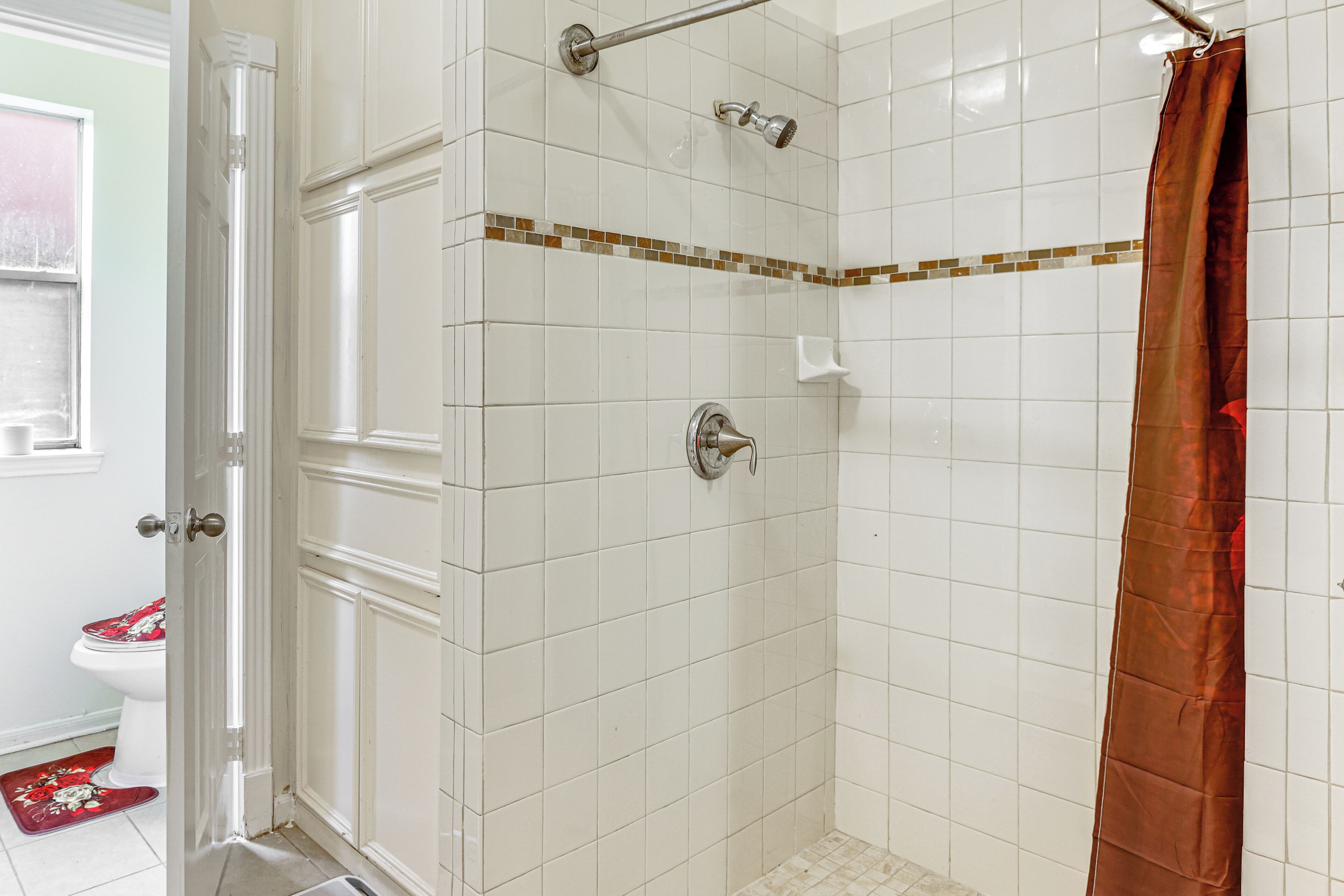 Casa de banho com paredes revestidas de azulejos brancos, chuveiro com cortina castanha e sanita com esteira vermelha.
