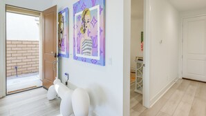 A hallway with a door leading to a balcony, a white wall with two framed artworks, and a white bench.