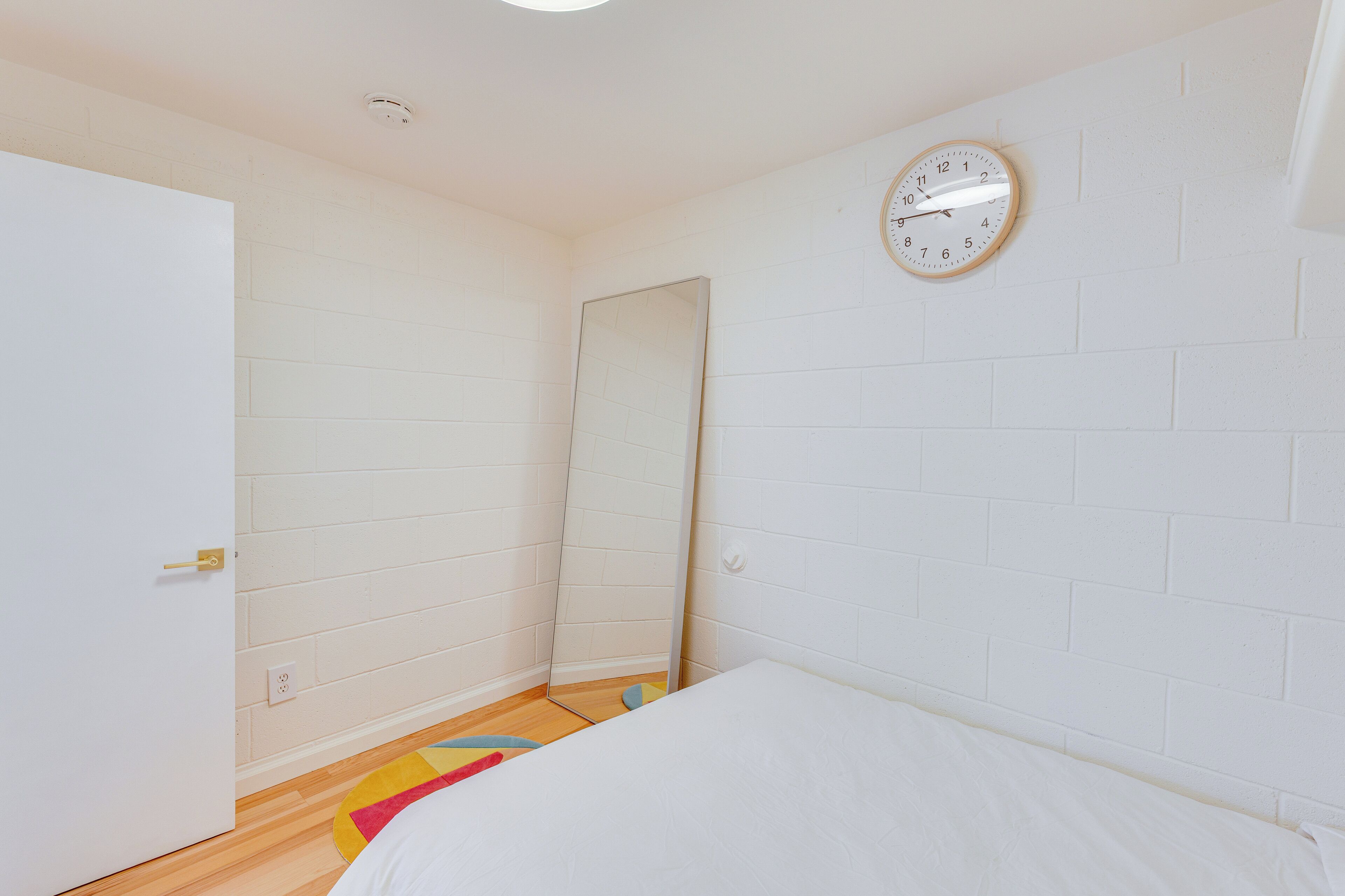 Una camera da letto con un letto, una porta bianca, uno specchio e una sveglia appesa al muro.