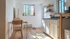 Une cuisine de taille réduite avec des meubles blancs, une chaise en bois, une petite table et une fenêtre.