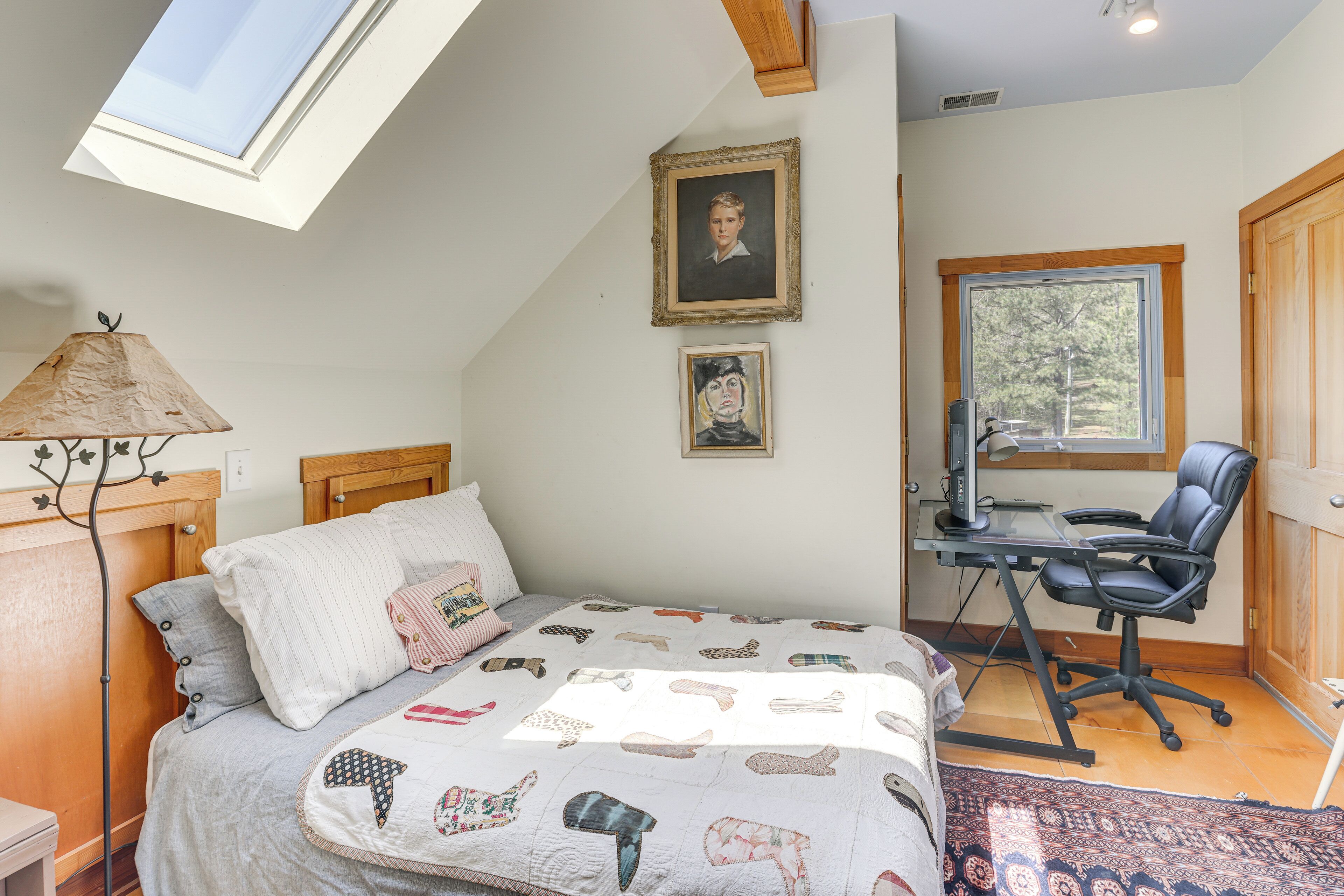 A bedroom with a sloped ceiling, a bed with a quilt, a desk with a chair, and a window with a view of trees.