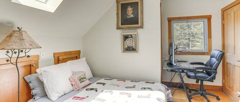 A bedroom with a sloped ceiling, a bed with a quilt, a desk with a chair, and a window with a view of trees.