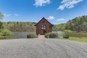 Une cabane en bois avec une allée en gravier, entourée d’un lac et d’une forêt.