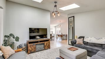 Uma sala de estar ampla com uma grande televisão de tela plana, sofás cinzas, um móvel para entretenimento de madeira e uma área de jantar ao fundo.